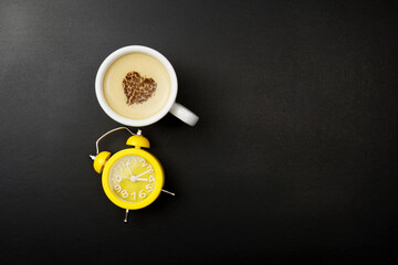 Cup of cappuccino and alarm clock, over black background with copy space. Chocolate heart in the center of a cup with a hot coffee drink and a yellow retro clock.