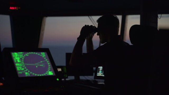 Captain looking through binoculars on ferry bridge during sunset, medium shot