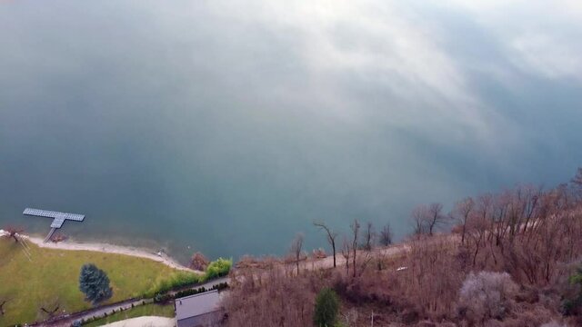Sweeping Drone aerial video footage above the Levico lake, facing the mountain Massiccio della vigolana, with sun setting in between the mountains behind the clouds. Flying above the water high