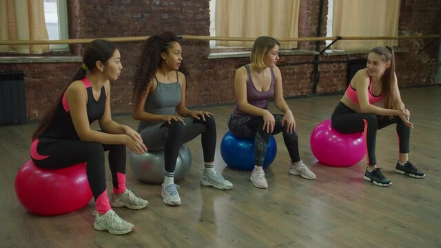 Positive relaxed lovely multiethnic diverse fit female friends in sports clothes resting on exercise balls, talking and sharing about pilates training class while taking a break during indoor workout.