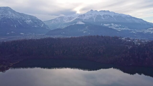Drone aerial video footage above the Levico lake, facing the mountain Massiccio della vigolana, with sun setting in between the mountains behind the clouds. Flying above the water