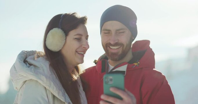 Caucasian adorable young married couple holding smartphone device taking picture on mobile phone wearing winter outfits outdoor on sunny weather.