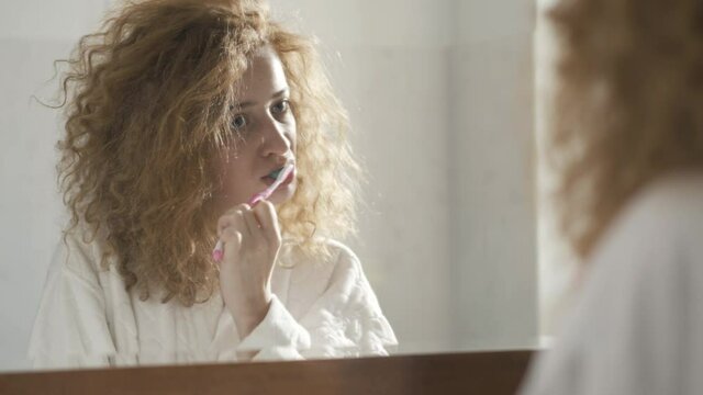 Close-up of confident woman brushing teeth reflecting in bathroom mirror. Portrait of adult redhead Caucasian lady in white bathrobe taking care of dentistry in the moning at home. Beauty and hygiene.