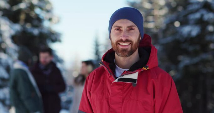 Portrait Of Candid Young Attractive Bearded Man In Winter Outfit Gathering With Friends Smiling To Camera Outside Snowy Forest Landscape.