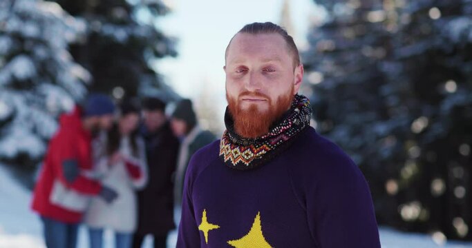 Nice handsome redheaded man with ginger beard in cozy sweater smiling at camera staying with friends on winter forest resort outdoors. Portraits.