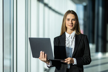 Portrait of young woman working holding laptop standing against panoramic window with city view