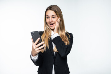 Fototapeta premium Portrait of surprised and impressed business woman with widened eyes and opened mouth, stunned on phone while standing against white background. Girl uses some app