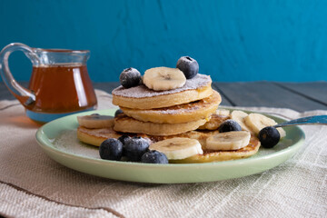 fresh pancakes with berries and chopped bananas on a plate, sprinkled with powdered sugar, next to a can of honey
