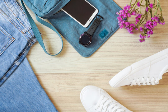 Top view or flat lay of faded blue jean , mobile phone , smart watch , hand bag , white sneaker and flower on wooden floor ,street trendy fashion accessories for girl or teenager 