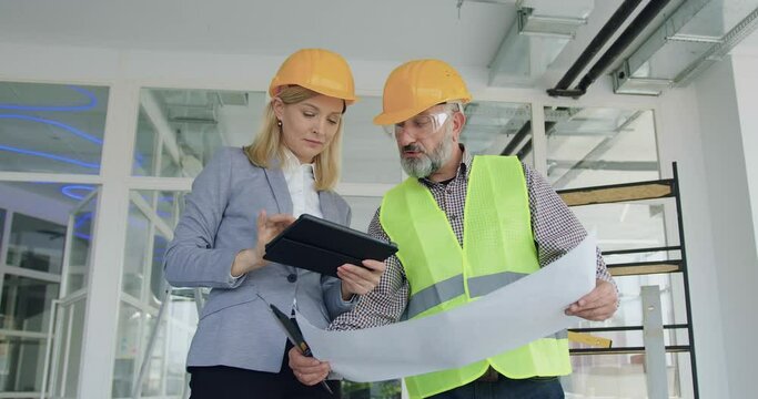 Beautiful purposeful skilled blond female manager in hardhat inspecting house or office together with professional elder bearded contractor and comparing all details according with plan on tablet