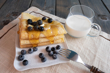 breakfast, blueberries, freshly baked pancakes poured with honey on a plate, next to a mug of milk