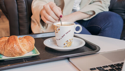 A working woman stirs the sugar in her coffee cup with a spoon accompanied by a croissant in a cafeteria. She eats breakfast while her computer is open for work. Morning routine 2021 during Covid 19.
