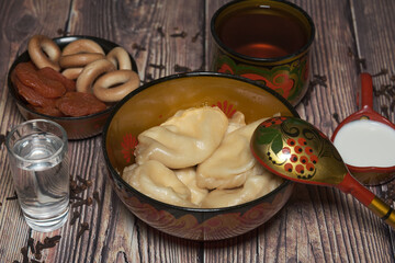 Russian Dinner in Khokhloma tableware. On the wooden table you can see Varenniki, bowl with dried apricots and bagels Sushki, sour cream, cup of herbal tea, scattered clove spices and glass of Vodka.