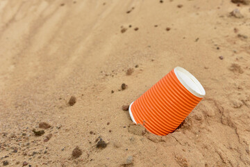 Fototapeta na wymiar Discarded Paper coffee cup on sand at beach. Disposable coffee cup on sandy background. The problem of environmental pollution. Pile of abandoned garbage, including food waste, fast food packaging