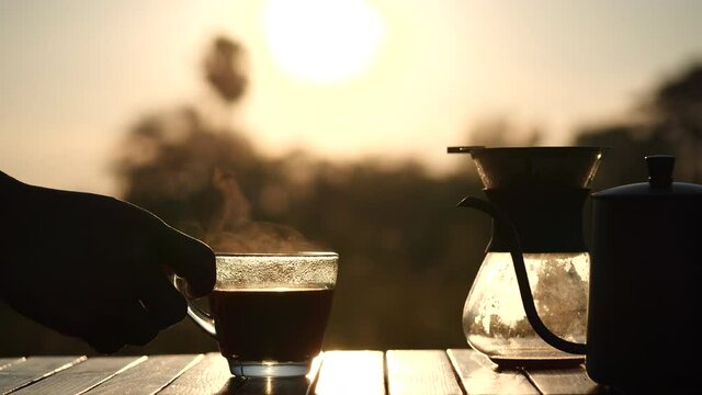 Men's hand takes a glass coffee cup with nature steam smoke of coffee on wooden, man drinks hot drip coffee while outdoor camping in beautiful nature sunrise background in the morning, slow motion.