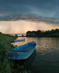 Storm arcus shaft and cumulonimbus cloud with heavy rain or summer shower, severe weather and sun glow behind rain. Landscape with Sava river with moored boats along grassy river bank and bridge.