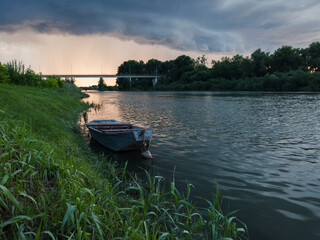 Storm arcus shaft and cumulonimbus cloud with heavy rain or summer shower, severe weather and sun glow behind rain. Landscape with Sava river with grassy bank, moored boat and bridge.