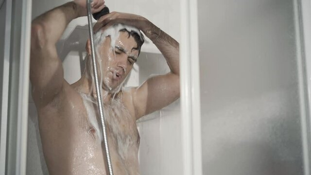 Handsome Caucasian man washing off shampoo in shower cabin. Portrait of confident brunette guy taking douche in the morning at home. Hygiene and routine concept.