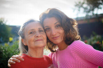 Senior mother with gray hair with her adult daughter looking at the camera in the garden and hugging each other during sunny day outdoors