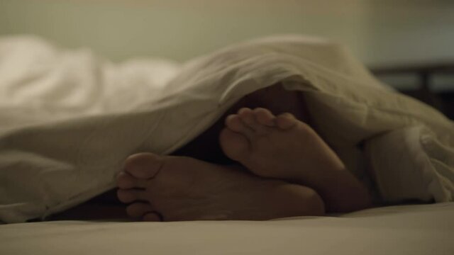 Male and female Caucasian feet stroking and hiding under blanket. Romantic adult couple flirting making out in bedroom at home. Intimacy and relationship concept.
