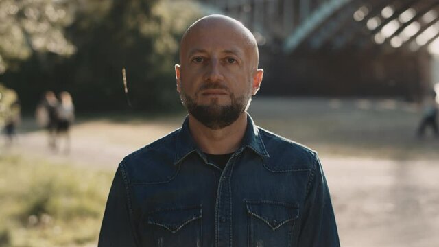 Face of bald hipster with a stylish beard is standing at the park. He is opening his eyes which were closed. His face has neutral expression.