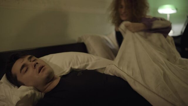 Angry stressed tired wife kicking out of bed husband snoring at night. Exhausted Caucasian woman pushing sleeping man. Marriage conflict and health care concept.