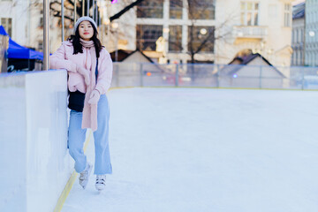 Full height of millennial of a young asian female wearing powder pink jacket and warm knitted hat, scarf and gloves skating on ice rink, outdoors at winter time.