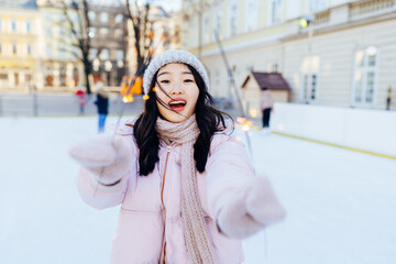 Obraz premium Inspired korean female with bengal lights with inspired smile spending vacations in european square to skate on rink. Asian woman in warm pink jacket, knitted hat, scarf, gloves in winter cold weather