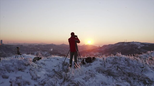 Panoramic view of a photographer with big camera standing on the hill and taking photos of bright winter sunset above Vladivostok. Russia