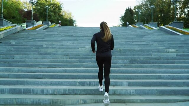 Woman with ponytail wearing black sportswear and white sneakers running upstairs in city park. Following shot of fit female jogging in the morning. Concept of fitness