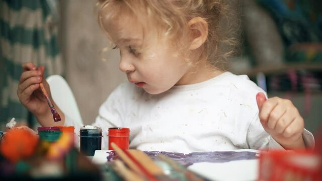 Blonde baby girl mixes tempera paints with a brush at home
