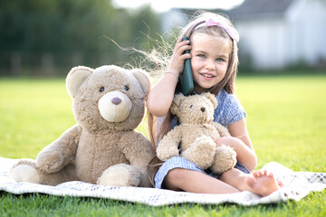 Pretty child girl sitting in summer park on green grass with her teddy bear toy talking on mobile phone outdoors in summer.