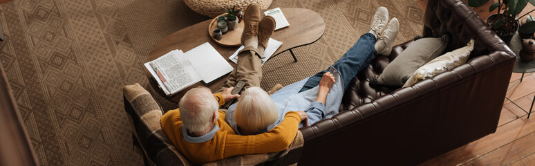 high angle view of elderly couple watching tv while hugging on couch in living room, banner