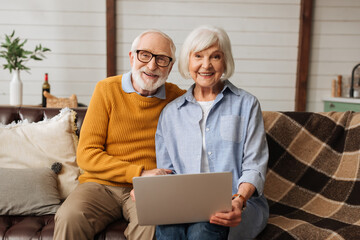 cheerful elderly husband hugging wife with laptop while looking at camera on couch in living room...