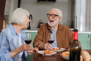 happy senior couple with wine glasses laughing while looking at each other at table with dinner on blurred foreground