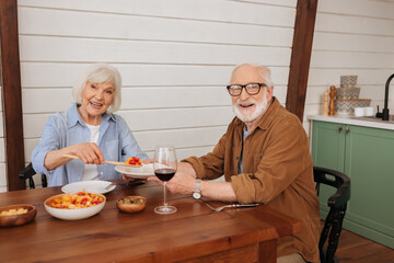 smiling senior wife looking at camera while serving salad for husband at table with vegetarian dinner in kitchen