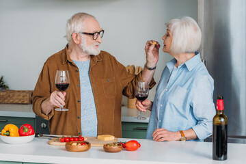 smiling senior man with wine glass feeding wife with piece of cheese in kitchen on blurred background