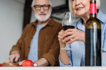 senior woman holding wine glass near bottle with blurred man on background indoors