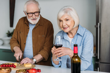 smiling senior husband looking at wife with wine glass while cutting cheese on chopping board in kitchen on blurred background