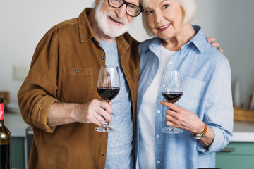 smiling senior couple hugging while holding wine glasses with blurred kitchen on background