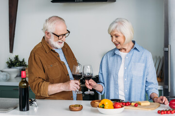 happy senior couple with wine glasses toasting near table with food in kitchen