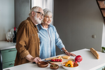 smiling elderly couple looking away while hugging near table with cheese, baguette and vegetables in kitchen