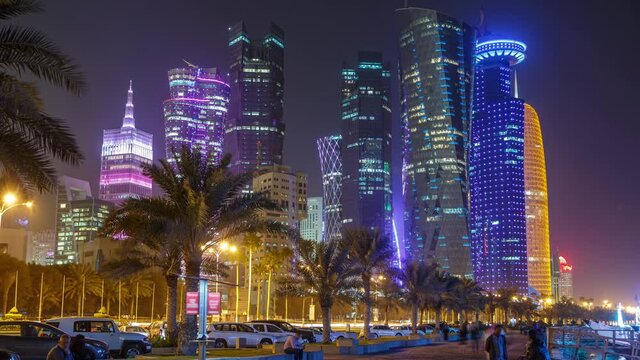 The skyline of Doha by night with walkway seen from Corniche timelapse, Qatar. Palms on foreground. Illuminated skyscrapers and towers