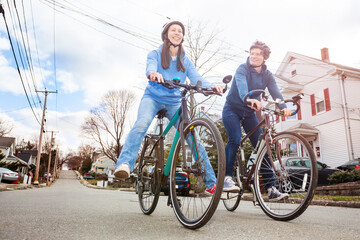 Fototapeta premium Low angle portrait of a laughing couple with smiling young woman ride a bicycle on the urban street wearing helmets