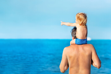 Fototapeta na wymiar Little toddler boy sit on shoulders of the father and point at the sea