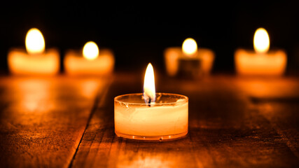 Candles in the dark on a table. Burning candle light closeup lit in the dark. Beautiful bokeh and tealight atmosphere glowing at night background. Christian faith and hope concept - obrazy, fototapety, plakaty