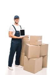 full length of cheerful mover in uniform and cap holding digital tablet with blank screen near carton boxes on white