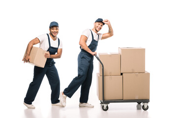 full length of positive multicultural movers in uniform and caps walking near hand truck with carton boxes on white