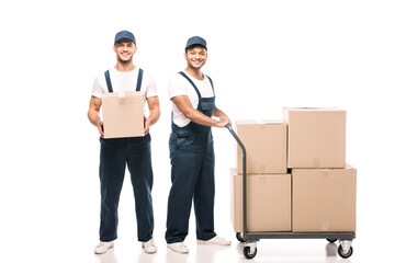 full length of happy multicultural movers in uniform and caps near hand truck with carton boxes on white