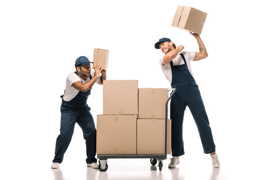 full length of angry tattooed mover in uniform holding carton box near scared indian coworker and hand truck with packages on white
